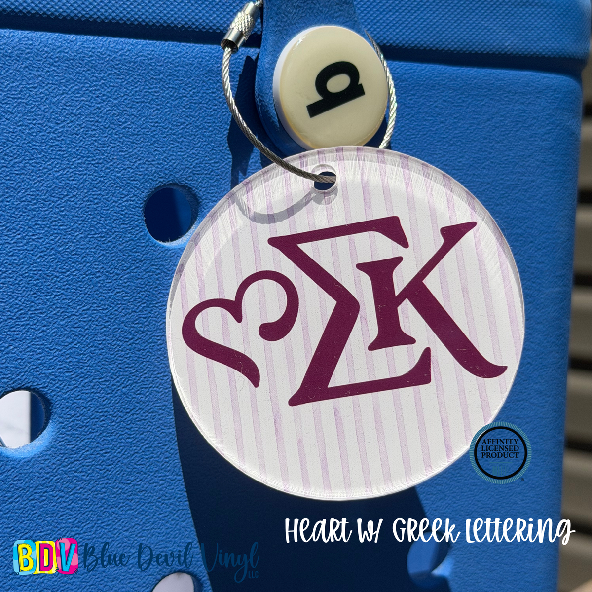 A round white acrylic keychain tag with a purple Greek letter monogram and a heart design, hanging from a silver key ring on a bright blue textured cooler. 
