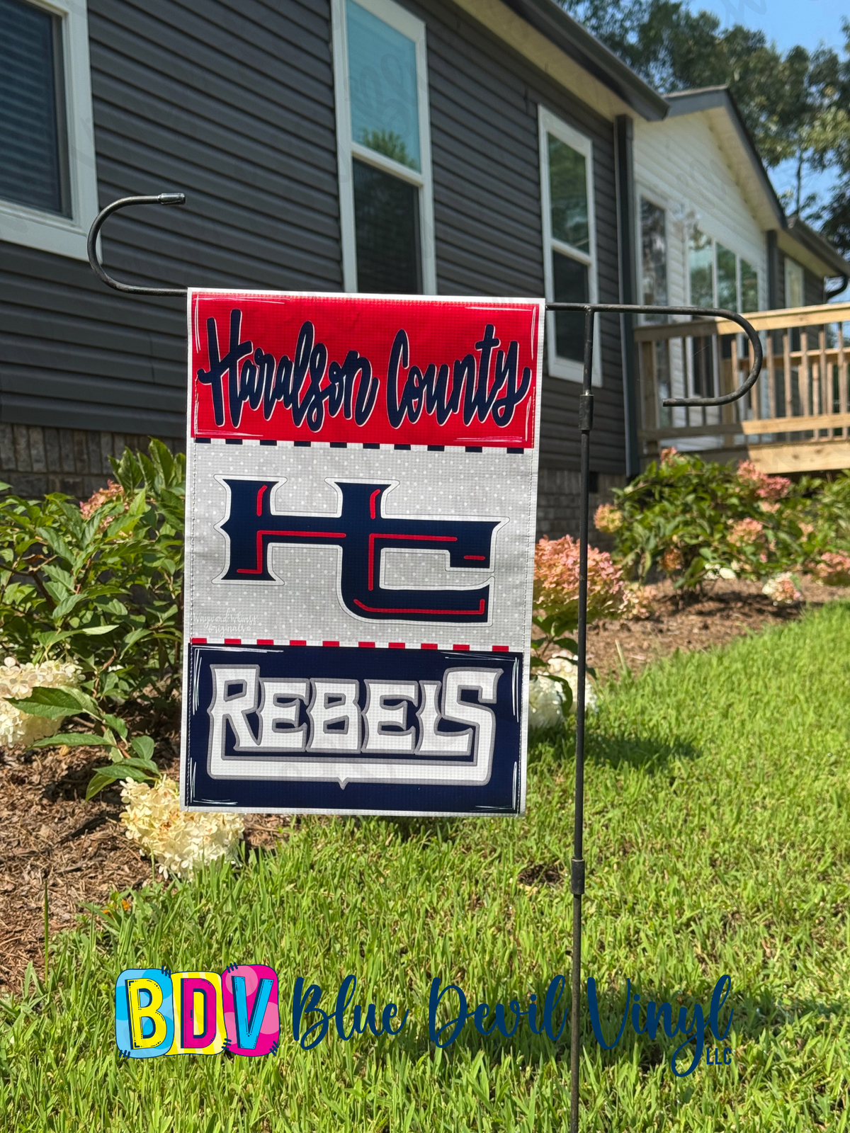 A rectangular yard sign with a red, white, and navy design reading 'Haralson County' at the top, a large 'HC' logo in the middle, and 'Rebels' at the bottom, displayed on a metal stand in a grassy yard in front of a house.
