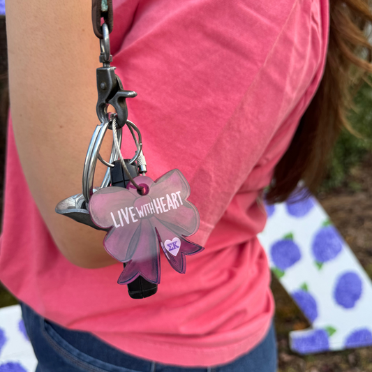 A close-up lifestyle shot of a person wearing a pink shirt, holding a keychain 
 that reads 'LIVE WITH HEART' in white text.