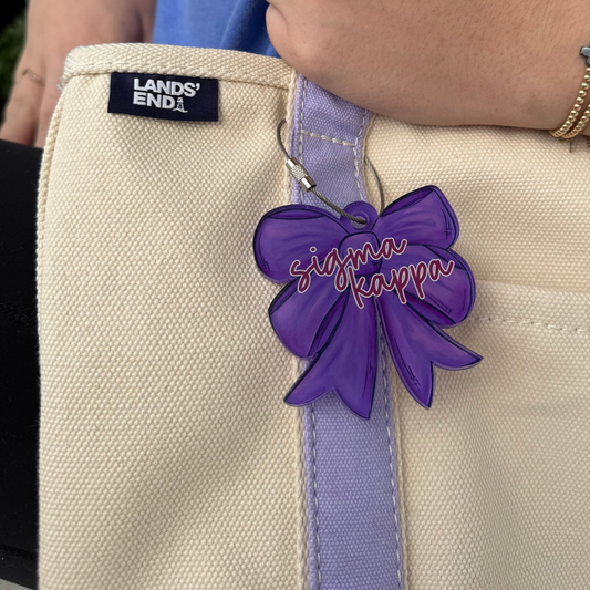 A close-up of a purple bow-shaped enamel pin with 'sigma kappa' written in pink script, hanging from a cream canvas bag with a lavender strap. 