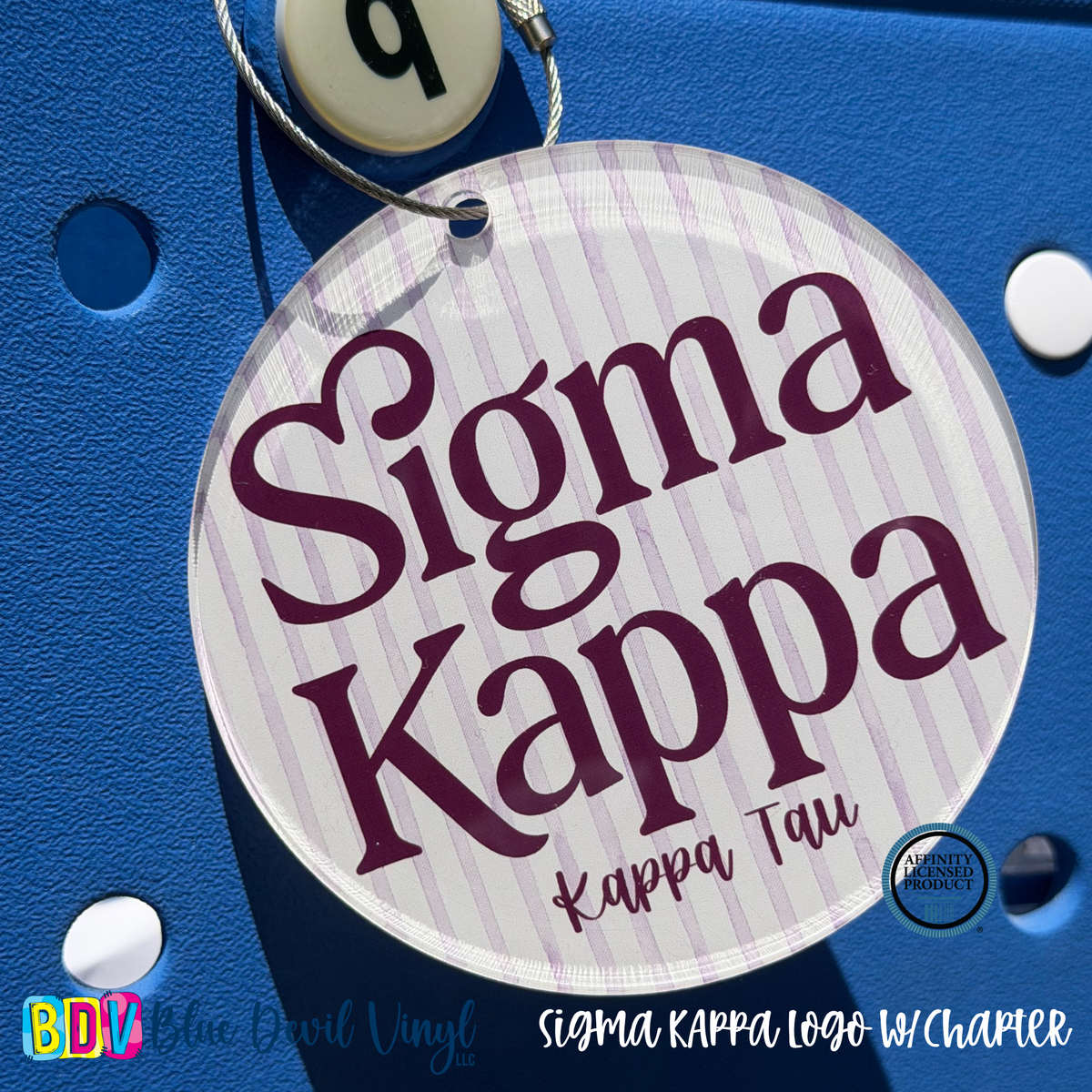 A round white and light purple striped Sigma Kappa keychain tag with the 'Sigma Kappa' logo in dark purple, shown hanging on a blue textured surface.